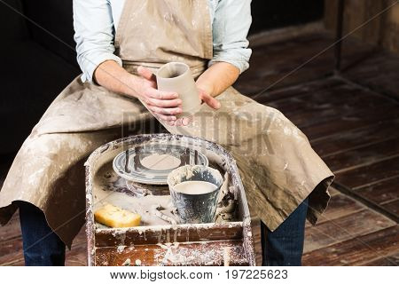 pottery, workshop, ceramics art concept - a master sits at the Potter's wheel, male hands hold unfired clay cup, a man examines a fresh product before further processing, front view