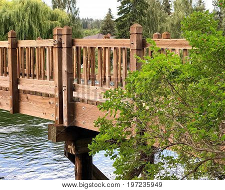 A wooden foot bridge with a berry bush spans the Deschutes River at Drake Park in Bend in Central Oregon on a summer day.