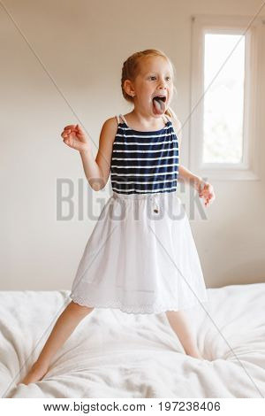 Portrait of cute finny white Caucasian blonde girl jumping on bed at home and showing tongue. Hilarious active kid having fun indoors. Authentic lifestyle childhood concept.