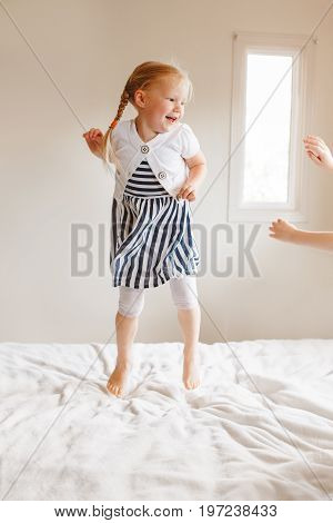Portrait of cute finny white Caucasian blonde girl jumping on bed at home and playing with sibling friend. Hilarious active kid having fun indoors. Authentic lifestyle childhood concept.