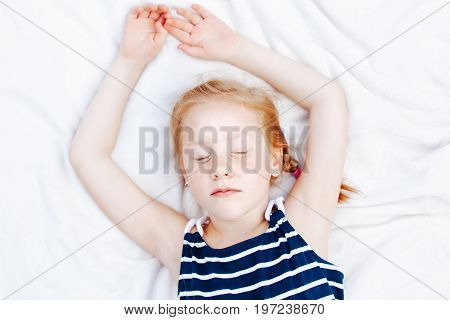 Closeup portrait of cute adorable redheaded Caucasian child girl in striped nautical sleeveless shirt sleeping. Kid dreaming lying on white blanket. Happy childhood dreams concept. View from above.