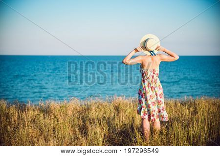 Summer Portrait Of Young Pretty Woman In A Straw Hat And Dress Standing Backwards In The Grass And L