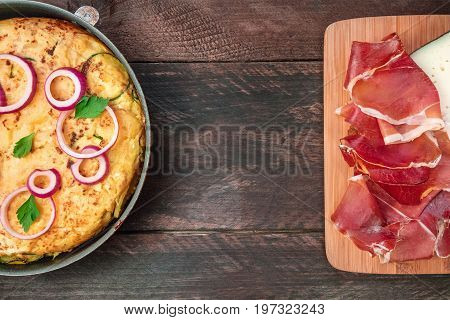 An overhead photo of a Spanish tortilla with potatoes and zucchinis in a tortillera, with jamon, cheese, and a place for text