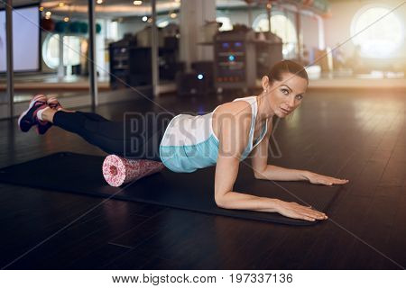 Woman in mint sleeveless shirt and black leggings in gym doing exercise with foam or fascia roll and looking at camera.