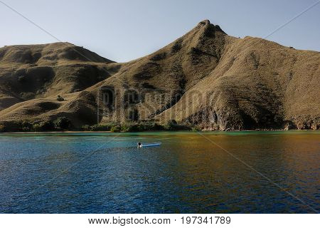 Small Boat In Front Of Coastline Of Mountains With Green Vegetation Reflected  In Blue Ocean Water A