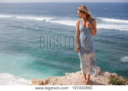Beautiful girl with hair floating in the air and light-blue dress is standing on the rock ledge in front of the ocean and looking away