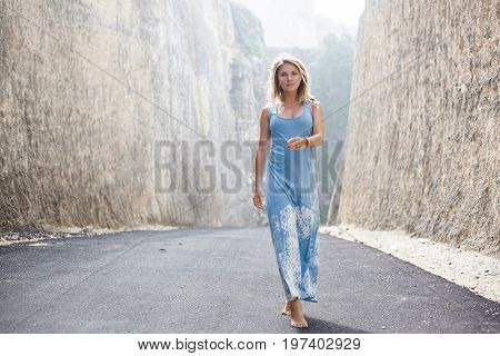 Attractive girl in light-blue dress is going forward on the road between mountains and looking in the camera