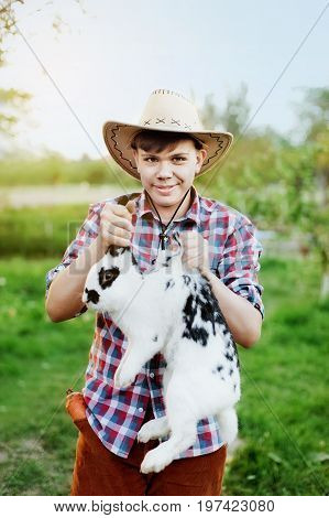 A boy in cowboy wear holding rabbit the scruff in farm. teenager holding a hare in the hands on the ranch at sunset