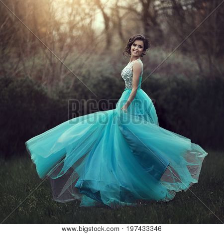 A beautiful graduate girl is spinning in a clearing in a blue dress. Elegant young woman in a beautiful dress in the park. Art photo.