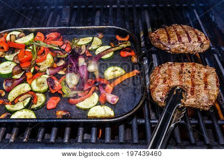 Two steaks being grilled on the barbecue along with roasted vegetables. The vegetables include zucchini red bell pepper green onions and mushrooms. The steak is being show about to be flipped with a pair of tongs.