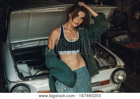 Girl worker in dirty green overalls and t-shirt stands in workshop near opened capote of retro car. Closeup.