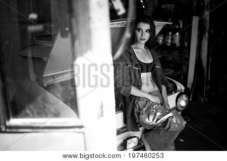 Girl worker in dirty overalls and t-shirt stands in workshop near opened capote of retro car. Black and white image.
