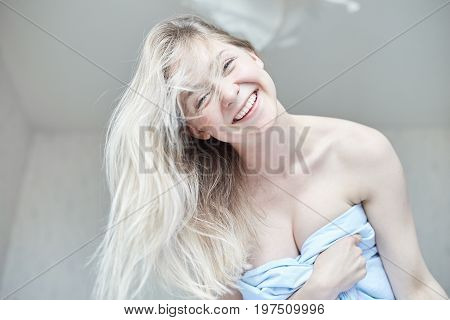 Tender indoor close-up portrait of beautiful Caucasian femaleposing with happy smile in her morning bed. Young attractive blue eyed lady with long blond hair looking playfully at the camera. Health and beauty concept.