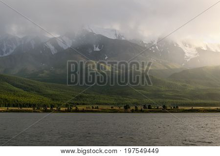 Scenic view with a lake mountains covered with snow forest cloudy sky fog and rain above the mountains with sunlight