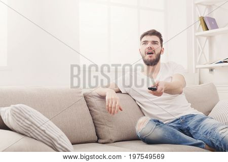 Young man wathing tv at home. Worried guy pointing with remote controller on tv-set, copy space