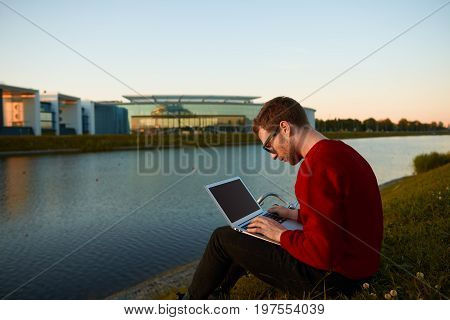 Outdoor shot of hardworking young European student in sunglasses studying remotely on notebook pc using free city wi-fi sitting on grass by the lake in public garden having concentrated look