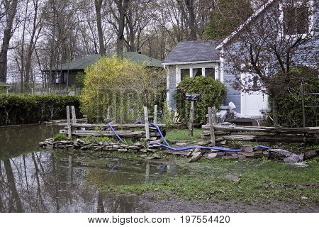Close up landscape view of flood waters rising over a paved road in front of properties with trees foliage and the houses partly submerged in the distance in the West Island of Montreal, Quebec on a bright overcast day in May.