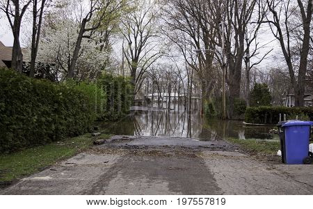 Landscape view of flood waters rising over a paved road in front of a gated property with trees foliage and a house partly submerged in the distance in the West Island of Montreal, Quebec on a bright overcast day in May.