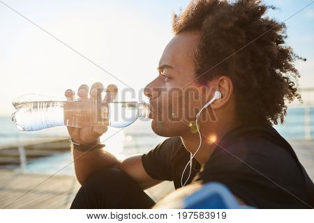 Outdoor shot of afro-american sportsman drinking water out of bottle in morning sunlight, wearing white earphones. Dark-skinned spotsman with bushy-hairstyle enjoying jogging in the morning by the sea. Healthy lifestyle concept.