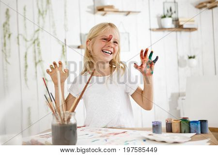 Smiling with teeth cute little blonde showing her hands in paint to the camera. Cheerful seven-year-old girl occupied with mess-free paint drawing.