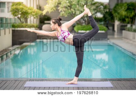Beautiful young Asian woman doing yoga exercise with Natarajasana posing (Lord of the dance pose) near swimming pool. Healthy lifestyle and good wellness concepts