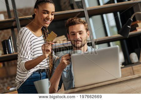Use my card. Positive man keeping smile on his face and wrinkling forehead while sitting close to his girl, looking at computer