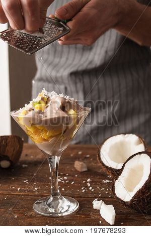 Culinary art of fresh fruit salad in martini glass with coconut flakes. Unrecognizable man makes a sweet dessert with decoration
