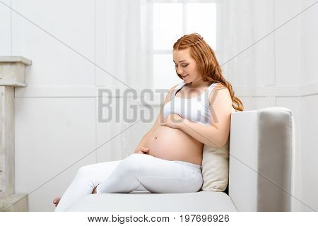 Happpy Beautiful Pregnant Woman Touching Her Belly While Sitting On Sofa At Home