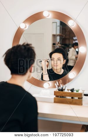 Attractive woman primp at the mirror with lamps in the beauty studio. A happy satisfied customer of make-up and hairstyle service.