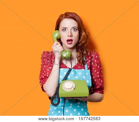 Surprised redhead girl in red polka dot dress and blue apron with green dial phone on yellow background.