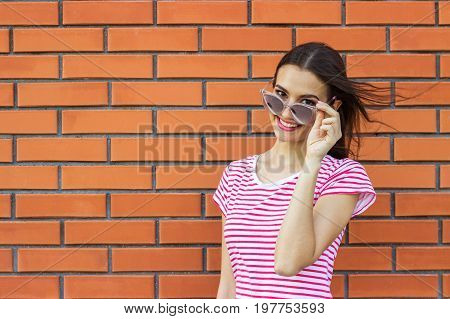 Close up portrait of cute girl wearing pink fashionable glasses looking aside over red brick wall background. Empty space for text design.