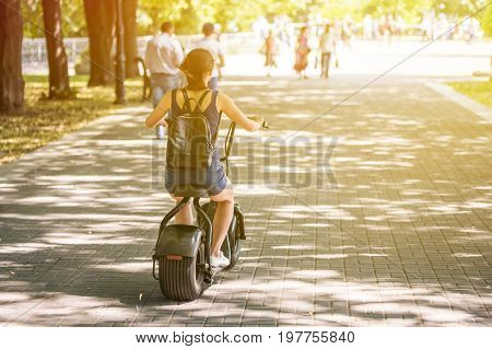 A young woman riding zero-emission eco electric scooter bike  in a city park. Back view. Day, sunlight.