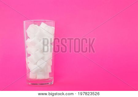A glass of sugar crystals or cubes on a bright pink background. Sweet food ingredient for cooking, baking, and beverages. Intake of bad calories.
