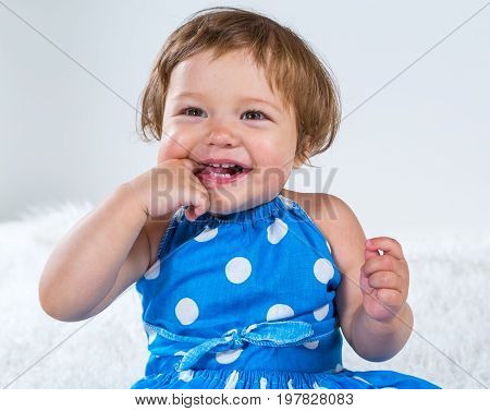 Little girl in a blue dress is sitting and laughing holding her finger in her mouth