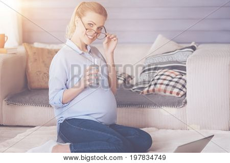 Source of energy. Cheerful happy pregnant woamn sitting on the floor and holding her glasses while drinking water