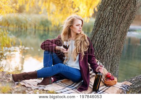 Beautiful woman drinking wine outdoors having picnic in the park. Portrait of young blonde beauty enjoying a glass of red wine sittind on autumn lawn near the lake