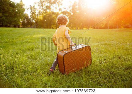 Curvy haired blonde kid wearing orange shirt carrying brown suitcase walking on grass.