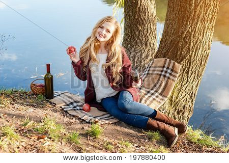 Beautiful woman drinking wine outdoors having picnic in the park. Portrait of young blonde beauty enjoying a glass of red wine sittind on autumn lawn near the lake