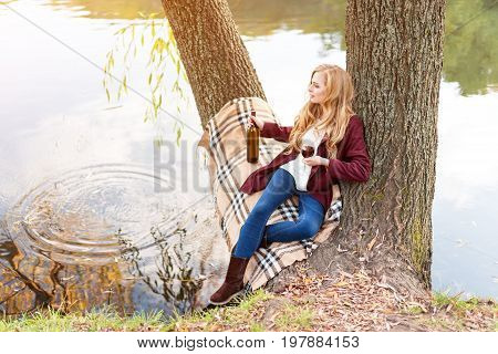 Beautiful woman drinking wine outdoors having picnic in the park. Portrait of young blonde beauty enjoying a glass of red wine sittind on autumn lawn near the lake