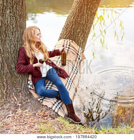 Beautiful woman drinking wine outdoors having picnic in the park. Portrait of young blonde beauty enjoying a glass of red wine sittind on autumn lawn near the lake