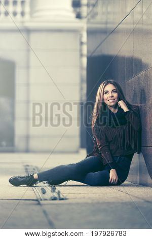 Happy young woman sitting at the wall in city street.  Stylish fashion model in leather fringe suede jacket and blue jeans