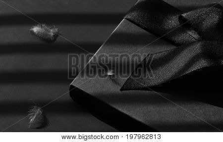 Black gift box on a dark contrasted background decorated with a textured bow and feathers creating a romantic atmosphere. Typically used for birthday anniversary presents gift cards post cards letters.