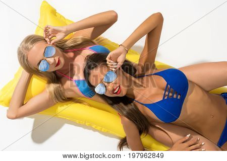 Sexy girls wearing sunglasses and bicinis lying on yellow swimming air mattress shot from above isolated