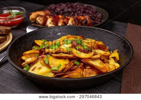Fried Potato Chips With Greens In A Pot, In Front Of Homemade Sausages And Stewed Cabbage, On A Blac