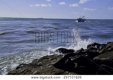 Waves Hitting The Rocks With Fishing Boat In The Background