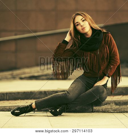 Happy young fashion woman sitting on city street sidewalk  Stylish female model in brown leather fringe suede jacket and black scarf