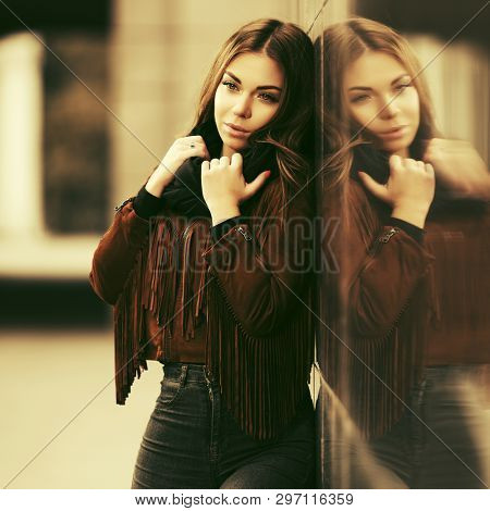 Sad young woman with long straight hairs leaning on the wall  Stylish fashion model in brown leather fringe suede jacket and black scarf