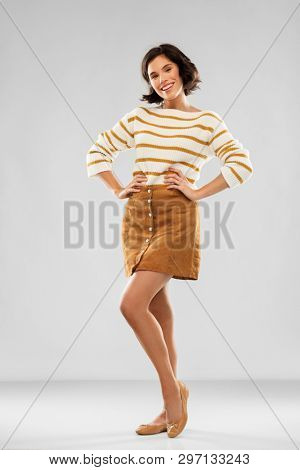people concept - happy smiling young woman in striped pullover, short skirt and ballet flat shoes with hands on hips posing over grey background