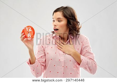 time, emotions and bedtime concept - shocked young woman in pajama with alarm clock showing half past six over grey background