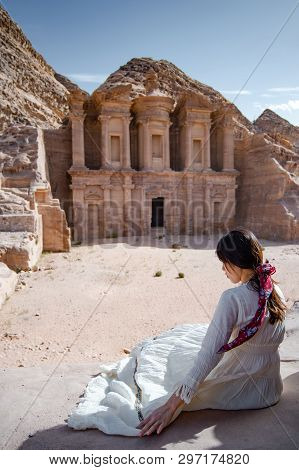 Asian Woman Tourist In White Dress Sitting At Ad Deir Or El Deir, The Monument Carved Out Of Rock In
